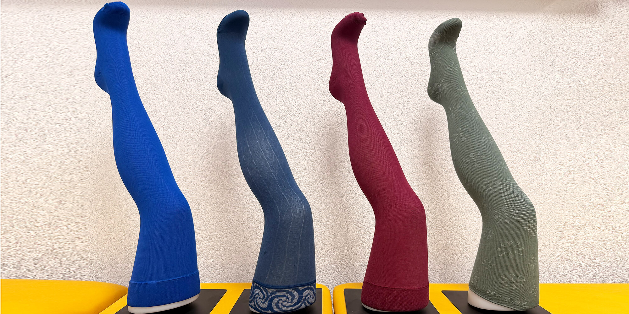 Detailansicht von Kompressionsstrümpfen in unterschiedlichen Farben mit Mitarbeitenden im Hintergrund einer Werkstatt
